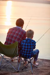 Papier Peint photo Pêcher Back view portrait of father and son sitting together on rocks fishing with rods in calm lake waters in sunset light, both wearing checkered shirts