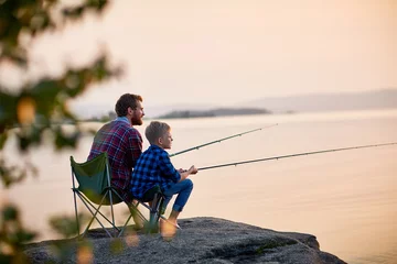 Foto op Plexiglas Vissen Zijaanzichtportret van vader en zoon die samen op rotsen zitten vissen met hengels in kalme meerwateren met landschap van ondergaande zon, beide met geruite shirts, geschoten van achter de boom