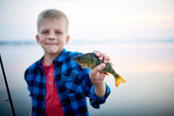 Portrait of teen blond boy smiling proudly holding one perch fish and showing it to camera against calm blue lake © pressmaster