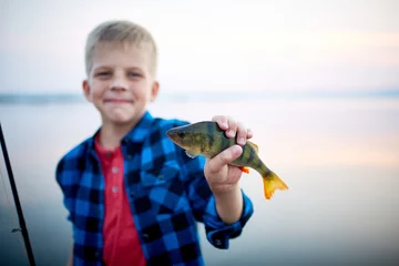 Tuinposter Portrait of teen blond boy smiling proudly holding one perch fish and showing it to camera against calm blue lake © pressmaster