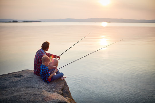 Back view portrait of father and son sitting together on rocks fishing with rods in calm lake waters with landscape of setting sun
