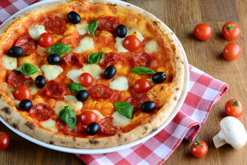 Hot tasty pizza with pepperoni, mozzarella, cherry tomatoes and basil, served on a wooden table for a dinner in italian restaurant. Pizza from wooden oven. Italy food concept.
