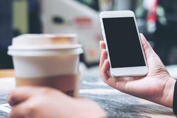Mockup image of hands holding white mobile phone with blank black screen and hot coffee cup in modern cafe