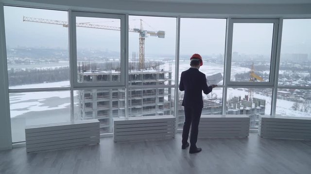 rear back view manager in contemporary company real estate property check documents and looking how growing house on construction site. elegant man in suit and hard hat in white modern develop office.