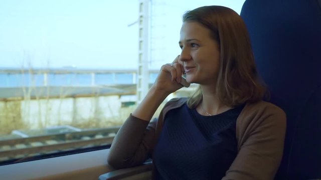 The girl is talking on the cell phone, sitting by the window in the train. In the window there are views of sea and city buildings