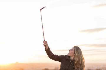 beautiful young woman taking a picture with a selfie stick at sunset. City background.
