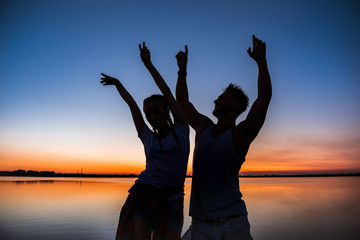 Fototapeta premium Silhouettes of young beautiful couple resting rejoicing at sunrise near lake.