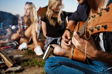 Friends sitting near bonfire, smiling, playing guitar. Camping grill marshmallow.