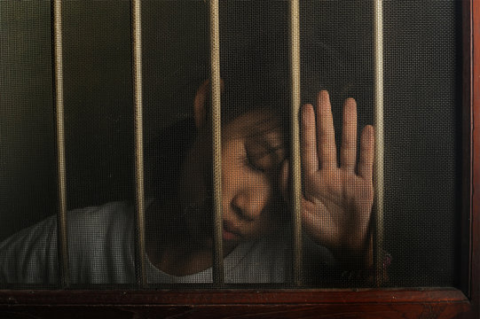 sad asian child standing behind the wire screen window in dark mood.  Unhappy child alone at home. Upset in the dark room