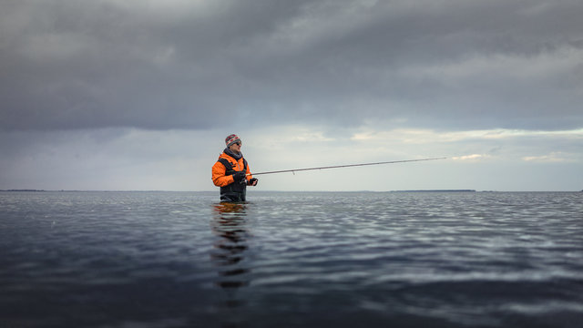 Angler mit Spinnrute watet in Wathose im Meer an der K&uuml;ste im kalten Winter bei Wolken