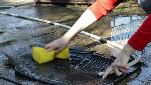 Washing rubber floor mats for car. Women's hand holding a yellow sponge and wash rubber floor mats for car with hose high pressure cleaner.