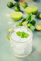 Ingredients for mojito cocktail, whole, sliced lime and mini limes, mint leaves, brown crystal sugar over gray stone texture background with glass full of ice. Space for text