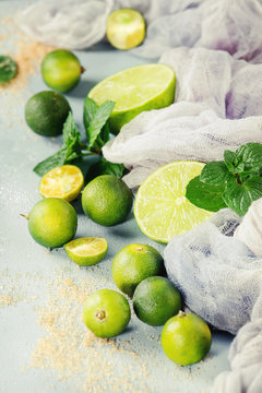 Ingredients for mojito cocktail, whole, sliced lime and mini limes, mint leaves, brown crystal sugar over gray stone texture background with gauze textile. Close up