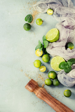 Ingredients for mojito cocktail, whole, sliced lime and mini limes, mint leaves, brown crystal sugar over gray stone texture background with gauze textile and wooden bar muddler. Top view, space