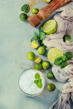Ingredients for mojito cocktail, whole, sliced lime and mini limes, mint leaves, brown crystal sugar over gray stone texture background, gauze textile, bar muddler, glass of ice. Top view, space