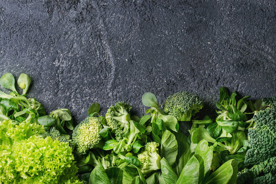 Variety of raw green vegetables salads, lettuce, bok choy, corn, broccoli, savoy cabbage as frame over black stone texture background. Top view, space for text