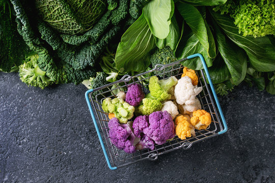 Variety of raw green vegetables salads, lettuce, bok choy, corn, broccoli, savoy cabbage, colorful young cauliflower in shop basket. Black stone texture background. Top view, space for text