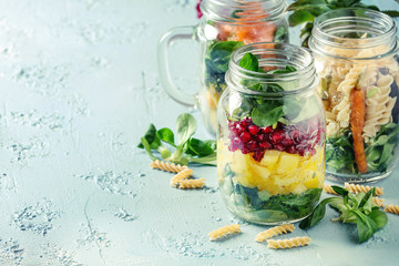 Variety of salads in mason jars. Fruit salad mango, pomegranate, greens, vegetables, wholegrain pasta, carrots and cauliflower, salmon. Standing over blue texture background. Food to go.