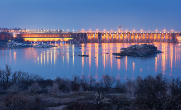 Dam at night. Beautiful industrial landscape with dam hydroelectric power station, bridge, river, city illumination reflected in water, rocks and sky. Dniper River, Zaporizhia, Ukraine.
