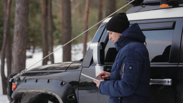 Lost driver looking around trying to find his way. Young man using his digital tablet to locate himself. In the background a pine forest and a car. Real time.