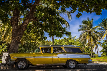 Amerikanischer gold gelber  Oldtimer parkt in Varadero  nahe des Strandes unter Palmen in Kuba - Serie Kuba Reportage