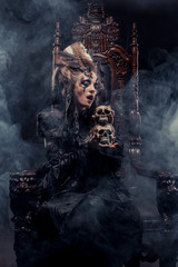 Young beautiful witch sits on a chair. Bright make up, skull, smoke-  halloween theme.