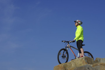 Fototapeta premium Man, in light-green jacket and red sneakers, near mountain bike with sky background