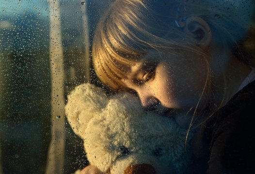 Sad child hugs a plush toy at rainy window.