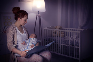 Young woman reading book for little baby in evening at home