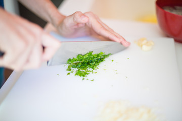 woman hands chopping green fresh parsley with big kitchen knife on white cutting board
