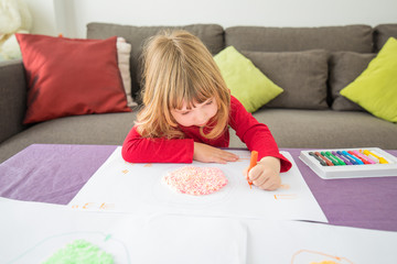 Three years blonde child red shirt with orange wax pencil or crayon in left hand drawing white sheet paper on table
