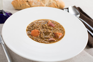 white elegant plate with lentils stew, orange carrot, ham and sausage, typical Spanish food, between silver knife, fork and spoon, brown cloth napkin, bread and crystal cup on beige tablecloth
