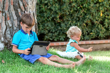 Two handsome happy boys and cute girl sitting with tablet on green grass