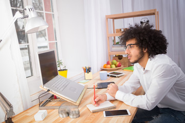 Hipster freelance man in glasses writing in notebook or diary and looking at laptop computer. Handsome man working at home alone.