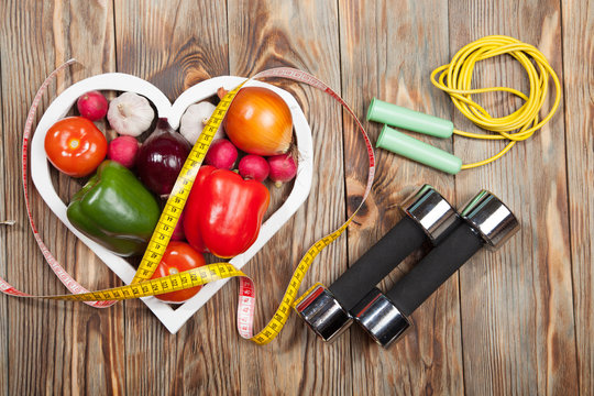 Sport and diet. Vegetables, dumbbells  centimeter. Peppers, tomatoes, garlic, onions,  radishes in the heart on rustic background