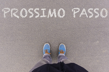 Fototapeta premium Prossimo passo, Italian text for Next Step text on asphalt ground, feet and shoes on floor