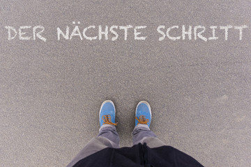 Der naechste Schritt, German text for Next Step text on asphalt ground, feet and shoes on floor