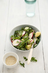 Spring salad from arugula, young potatoes, radish, cucumber and sesame seeds in ceramic bowl on light wooden table 
