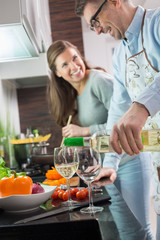 Man pouring white wine in glasses while cooking with woman at kitchen