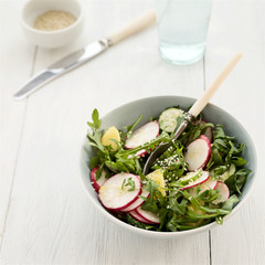 Spring salad from arugula, young potatoes, radish, cucumber and sesame seeds in ceramic bowl on light wooden table 
