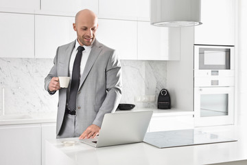 Mid adult businessman having coffee while using laptop in kitchen