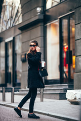 Fototapeta premium young stylish girl, passing by the Windows.Wearing fashionable glasses and a black coat.Keeps coffee.