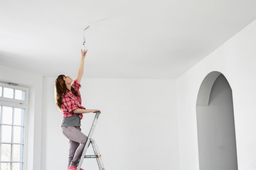 Full-length of woman on ladder fitting light bulb in new house