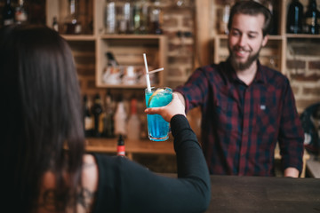Handsome bearded barman and beautiful young brunette woman toasting in cafe bar with blue lagoon cocktail.