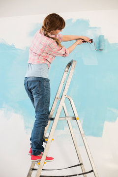 Woman On Ladder Painting Wall With Paint Roller