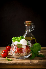 Delicious caprese salad with quinoa, ripe cherry tomatoes and mini mozzarella cheese with fresh basil leaves in glass jar and olive oil. Italian healthy food concept with copy space. Gluten free.
