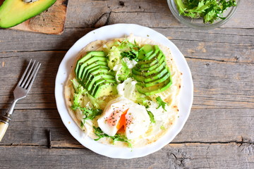 Vegetarian breakfast with a poached egg, avocado slices, chinese cabbage, lettuce, tortilla, sauce and spices. Easy avocado and poached egg tortilla on a plate and on old wooden background. Top view