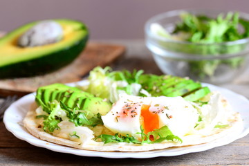 Healthy breakfast with a poached egg, avocado slices, chinese cabbage, lettuce, tortilla, sauce and spices. Simple avocado and poached egg tortilla on a plate and on old wooden background. Closeup