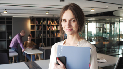 portrait of positive attractive brunet young working woman who look and smile to camera holding blue folder with business papers staying on front of working people in light office with big windows