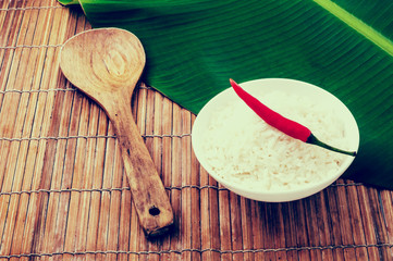 White bowl with Steam cooked white Jasmine rice and red chili pepper on top. Asian countryside background with banana leaf and bamboo table cover with wood spoon. Retro color fade out effect